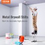 worker on VEVOR drywall stilts painting a ceiling, surrounded by paint supplies in a white room.