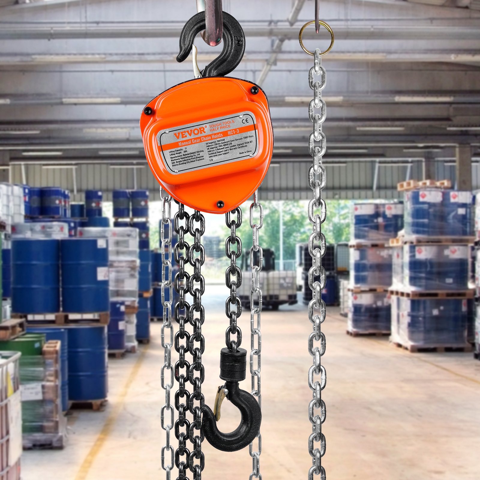 VEVOR hand chain hoist in an industrial warehouse with barrels and pallets in the background.