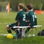 soccer players seated on a VEVOR folding bench with a yellow ball on the grass.