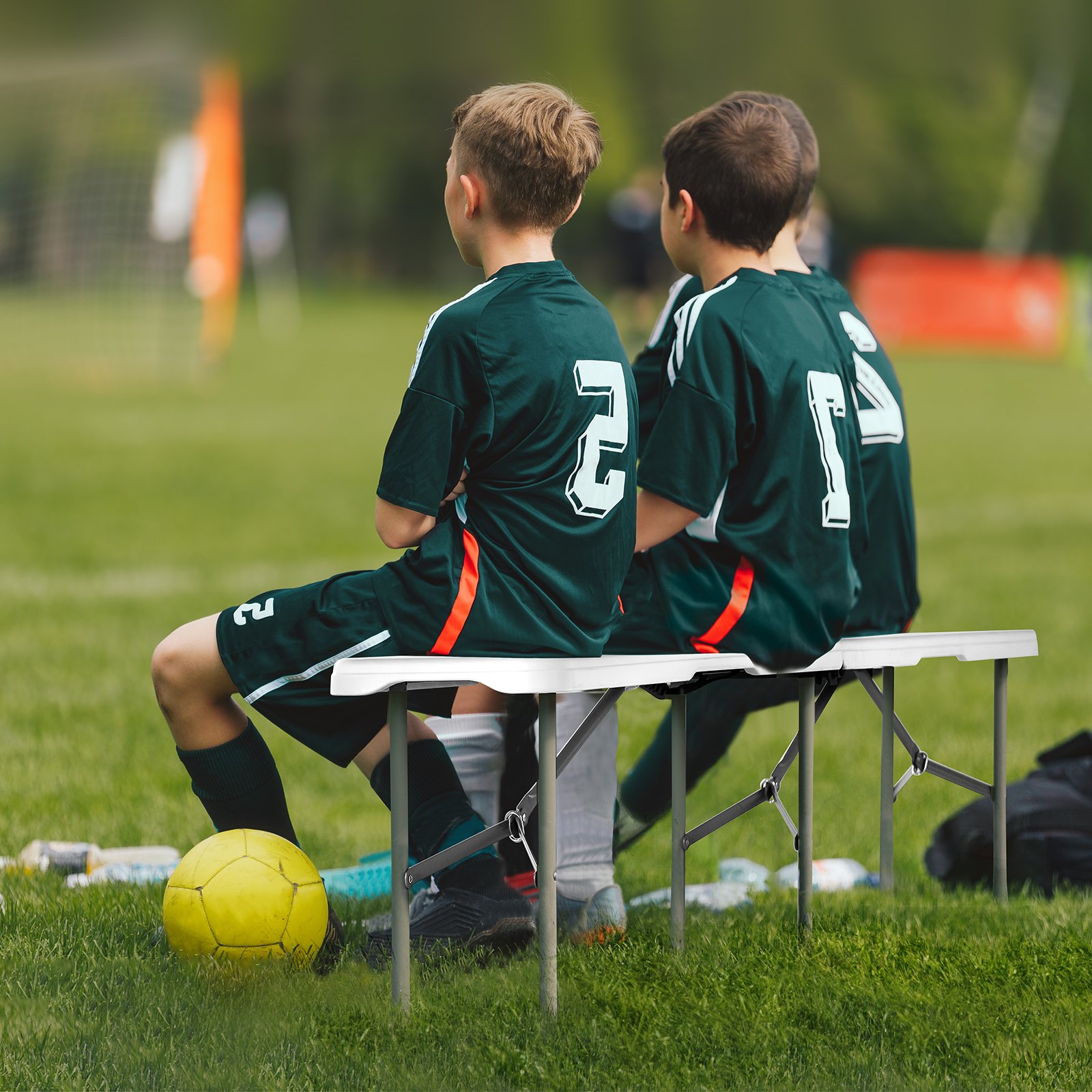 soccer players seated on a VEVOR folding bench with a yellow ball on the grass.