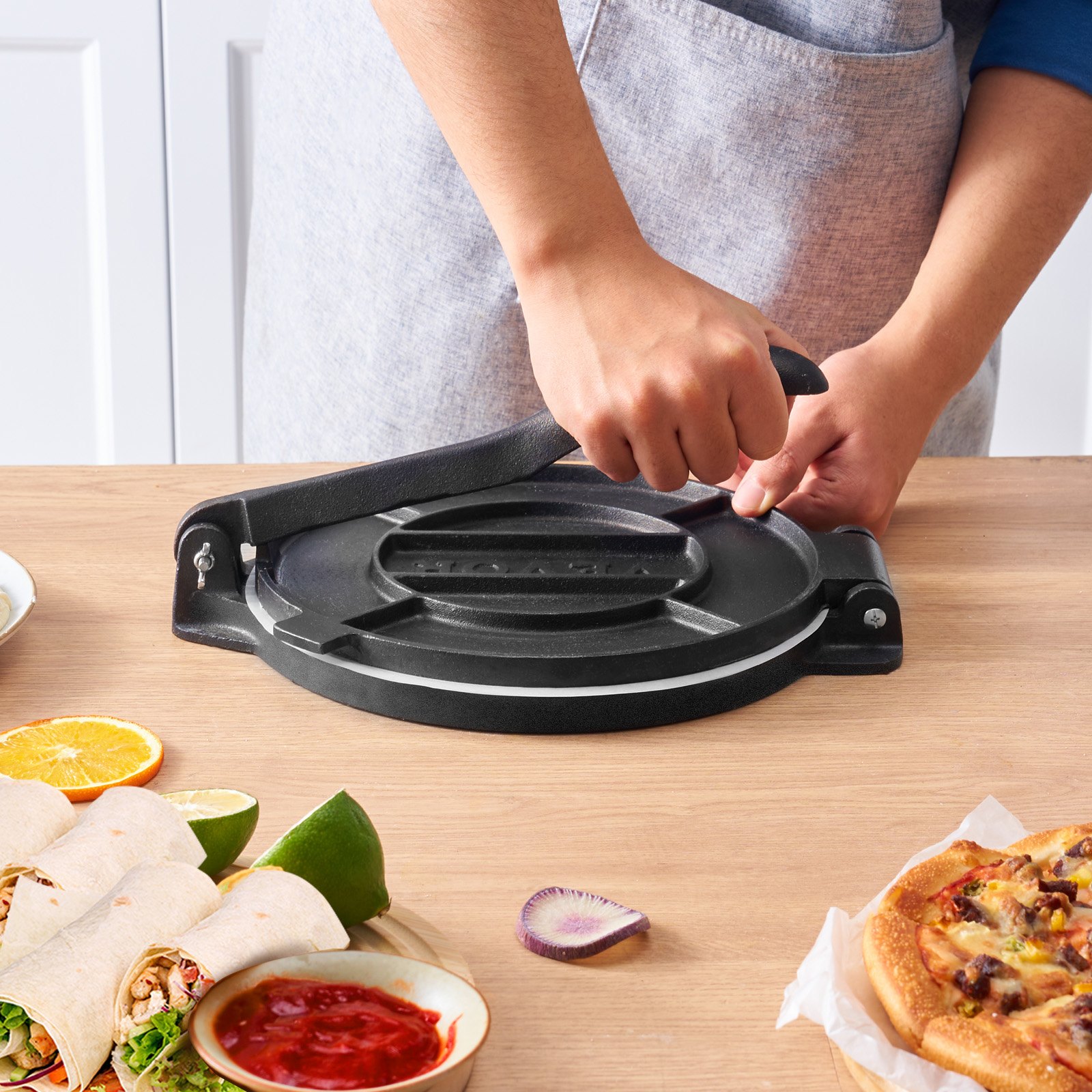 person using a VEVOR tortilla press on a wooden table with wraps, salsa, and pizza nearby.