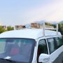 white van equipped with a VEVOR van roof ladder rack carrying lumber and pipes, with greenery in the background.