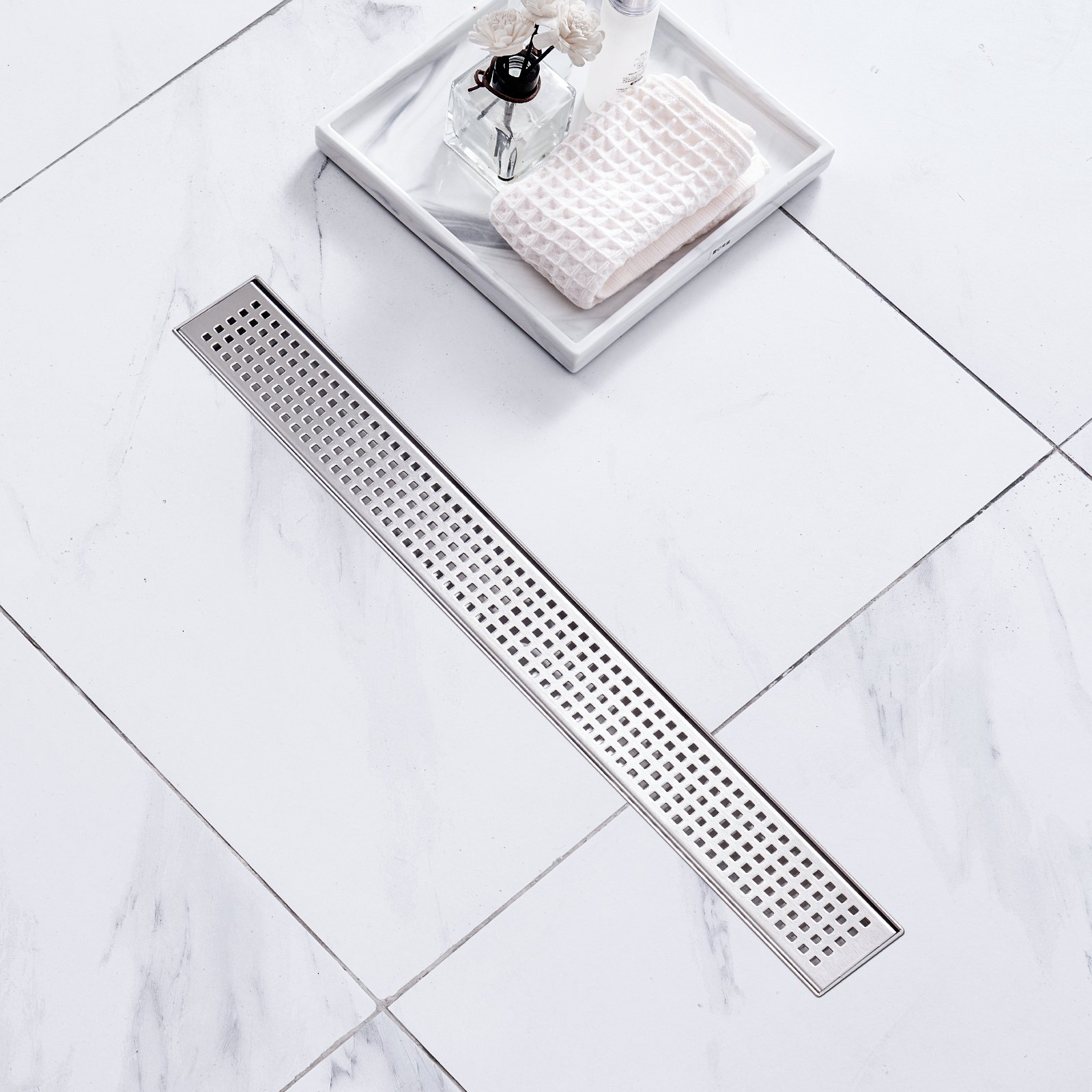 VEVOR linear shower drain on white marble tile floor with a tray, soap, and flowers nearby.