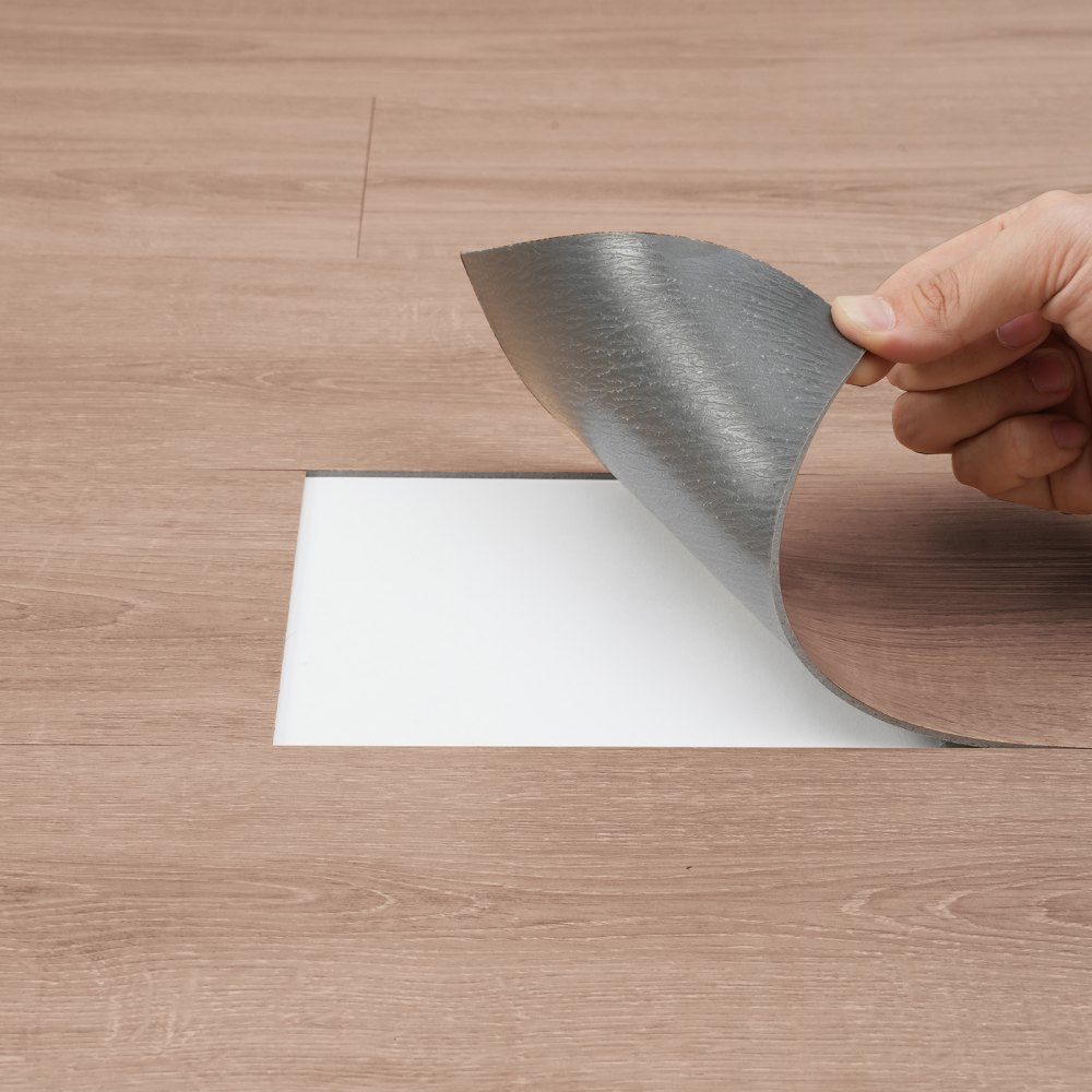 hand installing VEVOR self adhesive vinyl floor tiles on wood-patterned surface.